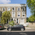 Three-story brick townhouse with arched white-trimmed windows, parked cars on the street, and leafy trees on a sunny day.