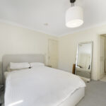 Bedroom with white bedding and a gray upholstered headboard, a black office chair, and a wooden side table; a large standing mirror leans on the wall and an open bathroom door is visible.