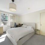 Bright hotel-like bedroom with a large white bed, cream walls, and a window letting in sunlight; desk chair and small side table nearby.