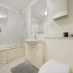 White bathroom with a bathtub, glass shower enclosure, and a white sink and vanity. A large mirrored cabinet and light wash walls fill the space, with a gray rug on the floor.