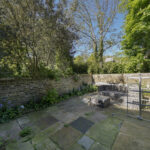 Sunny backyard garden with a stone-paved patio, wicker seating area, and a brick retaining wall framed by tall trees and greenery under a blue sky.