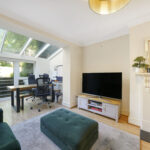 Bright living room with a green sofa and ottoman, a fireplace on the right, and a TV cabinet; an open entry leads to a glass-walled conservatory workspace beyond.