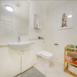 White bathroom with pedestal sink, mirror cabinet, and toilet; wall art of a standing otter holding a towel above a wooden shelf of toiletries on the right.
