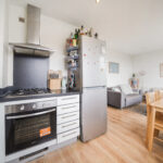 Modern kitchen with stainless steel appliances, including a tall fridge and gas range, adjacent to a living area with wall art and a wooden dining table.