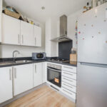 Modern L-shaped kitchen with white cabinets, black countertop, stainless steel range hood, oven, and a large silver fridge with magnets on the door. A microwave sits on the counter beside a dish rack and sink on the left side.