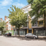Tree-lined urban street with modern brick buildings, parked cars, and a bright blue sky with a few clouds.