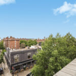 Urban street scene with brick buildings, parked cars, and trees under a clear blue sky, viewed from a rooftop railing.