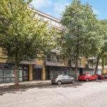 Tree-lined urban street with modern brick buildings, glass storefronts, and parked cars in front.