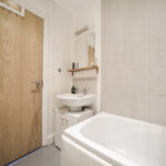 Narrow bathroom with a wooden door on the left, white sink and cabinet, mirror, and tiled wall on the right.”