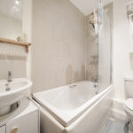 White bathroom with a sink and vanity on the left, a bathtub with a glass shower screen in the middle, and a toilet on the right.