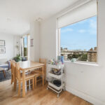Bright living area with a wooden dining table and chairs, a gray sofa with cushions, and framed wall art beneath a large window showing a city view.