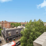 Urban street scene viewed from a high balcony: brick buildings, storefronts, parked cars, and green trees under a clear blue sky.