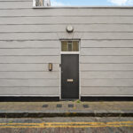 Grey exterior wall with a centered dark door and small window above, brick side wall on left, street and curb in foreground.