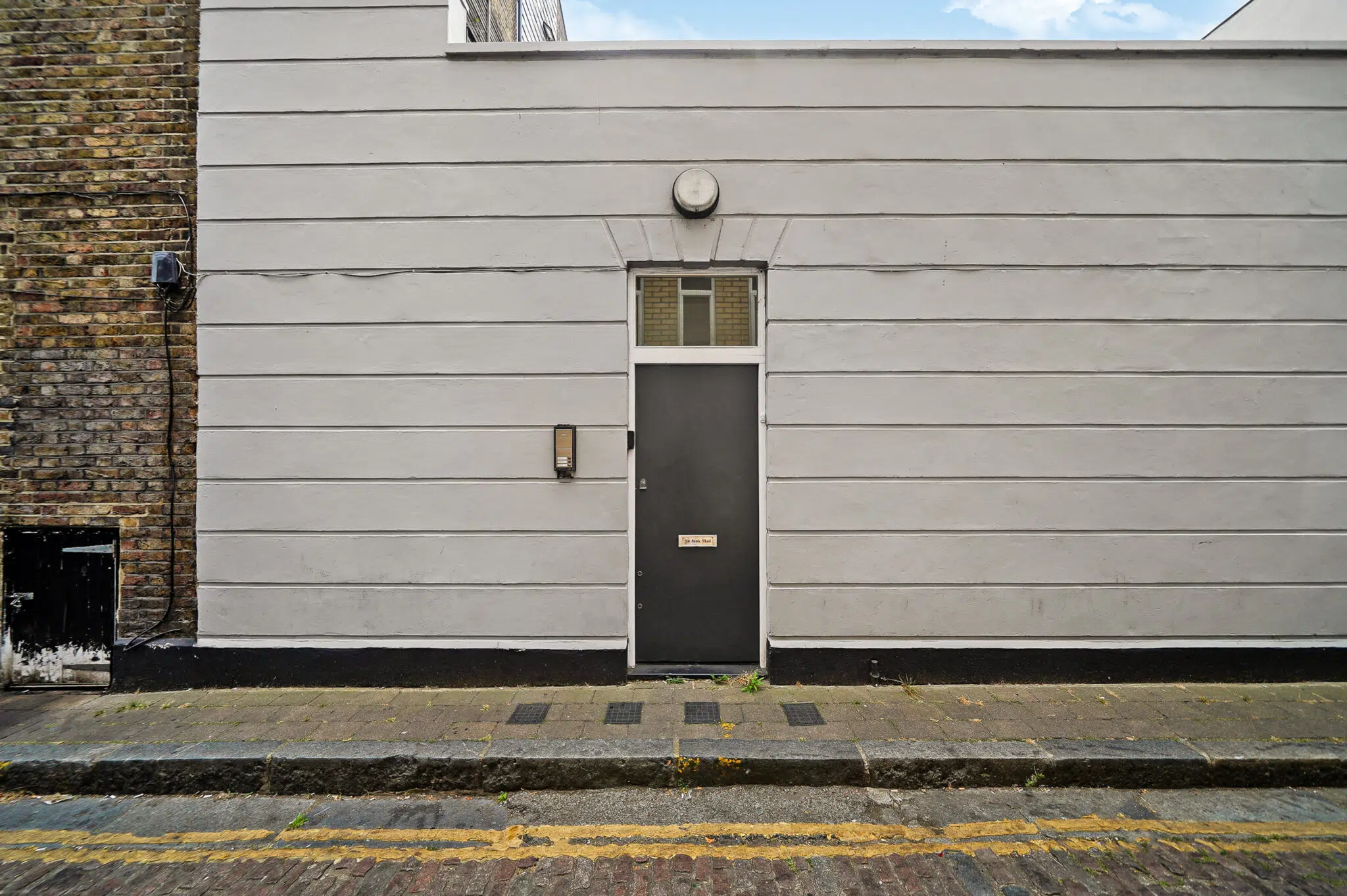Grey exterior wall with a centered dark door and small window above, brick side wall on left, street and curb in foreground.