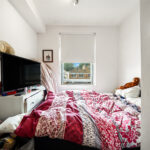 Bedroom scene: unmade bed with a red and white quilt, a window at the foot of the bed, and a TV on a white dresser with a standing fan nearby.