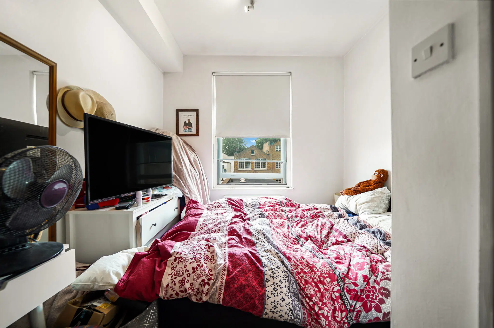 Bedroom scene: unmade bed with a red and white quilt, a window at the foot of the bed, and a TV on a white dresser with a standing fan nearby.