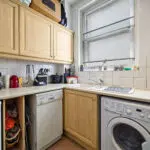 Compact kitchen corner with light wood cabinets, kettle and red canister on the counter, and a front-loading washing machine beneath the counter next to a sink.