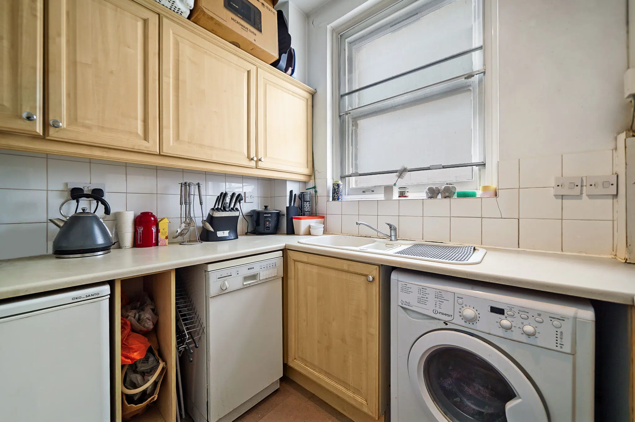 Compact kitchen corner with light wood cabinets, kettle and red canister on the counter, and a front-loading washing machine beneath the counter next to a sink.