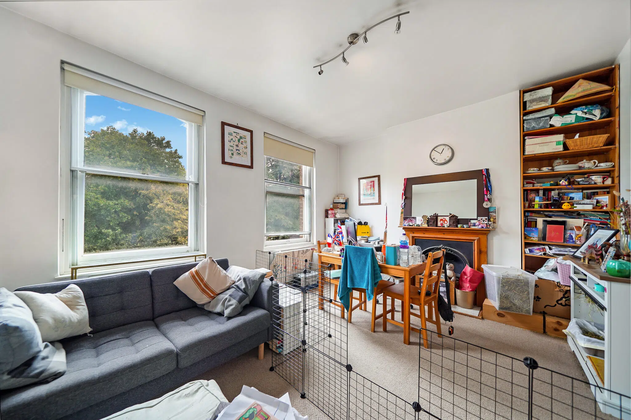 Living room with two large windows, a gray sofa, and a metal pet pen in the center amid cluttered shelves and table items outside the windows.