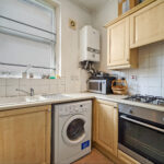 Compact kitchen with a wall-mounted boiler, washing machine under the counter, and a gas range next to wooden cabinets across tiled walls and a window above the sink