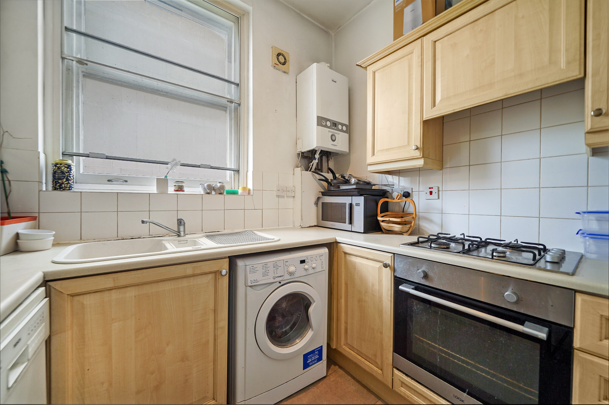 Compact kitchen with a wall-mounted boiler, washing machine under the counter, and a gas range next to wooden cabinets across tiled walls and a window above the sink