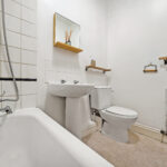 White bathroom with a bathtub in the foreground, sink and toilet along the wall, and a large radiator on the right side; wooden shelves and a mirror above the sink add storage.