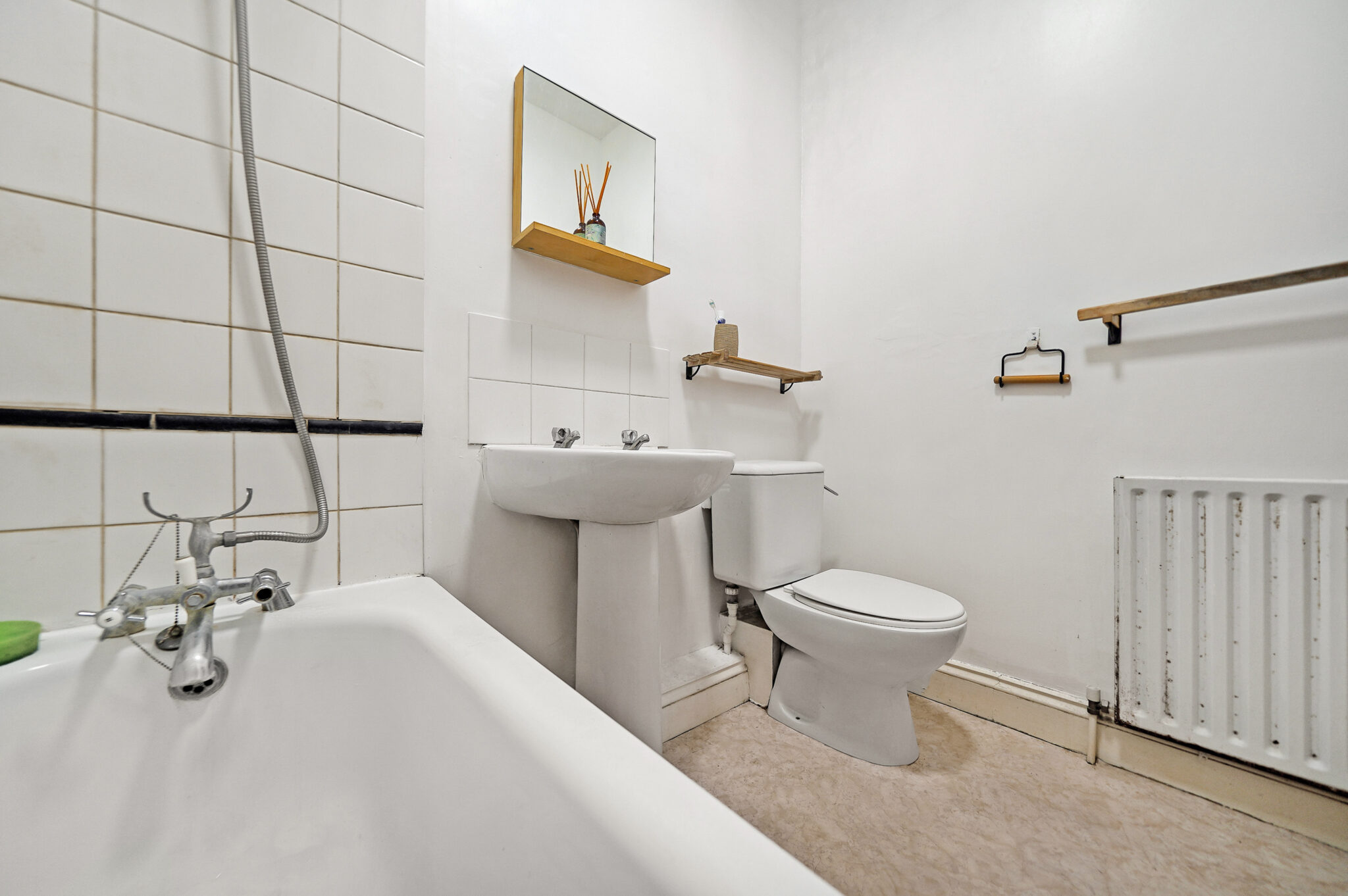 White bathroom with a bathtub in the foreground, sink and toilet along the wall, and a large radiator on the right side; wooden shelves and a mirror above the sink add storage.