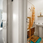 View from a doorway into a bathroom with a white pedestal sink, orange shower curtain, and teal rug on a wooden slat mat.