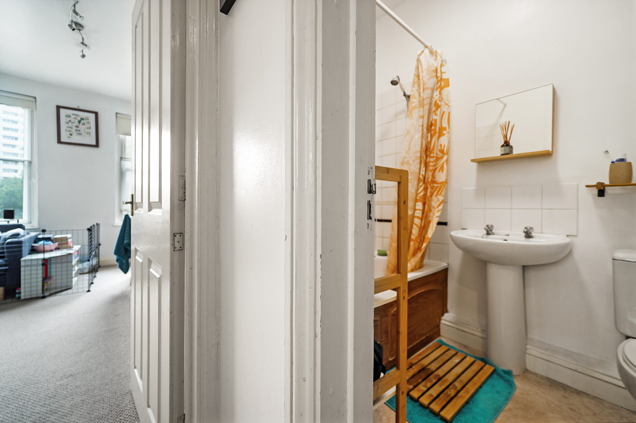 View from a doorway into a bathroom with a white pedestal sink, orange shower curtain, and teal rug on a wooden slat mat.