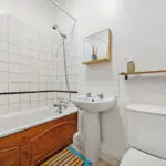 Compact bathroom with white tile walls, a wooden-front bathtub, pedestal sink, and a toilet. A wall mirror shelf and towel bar are also visible.