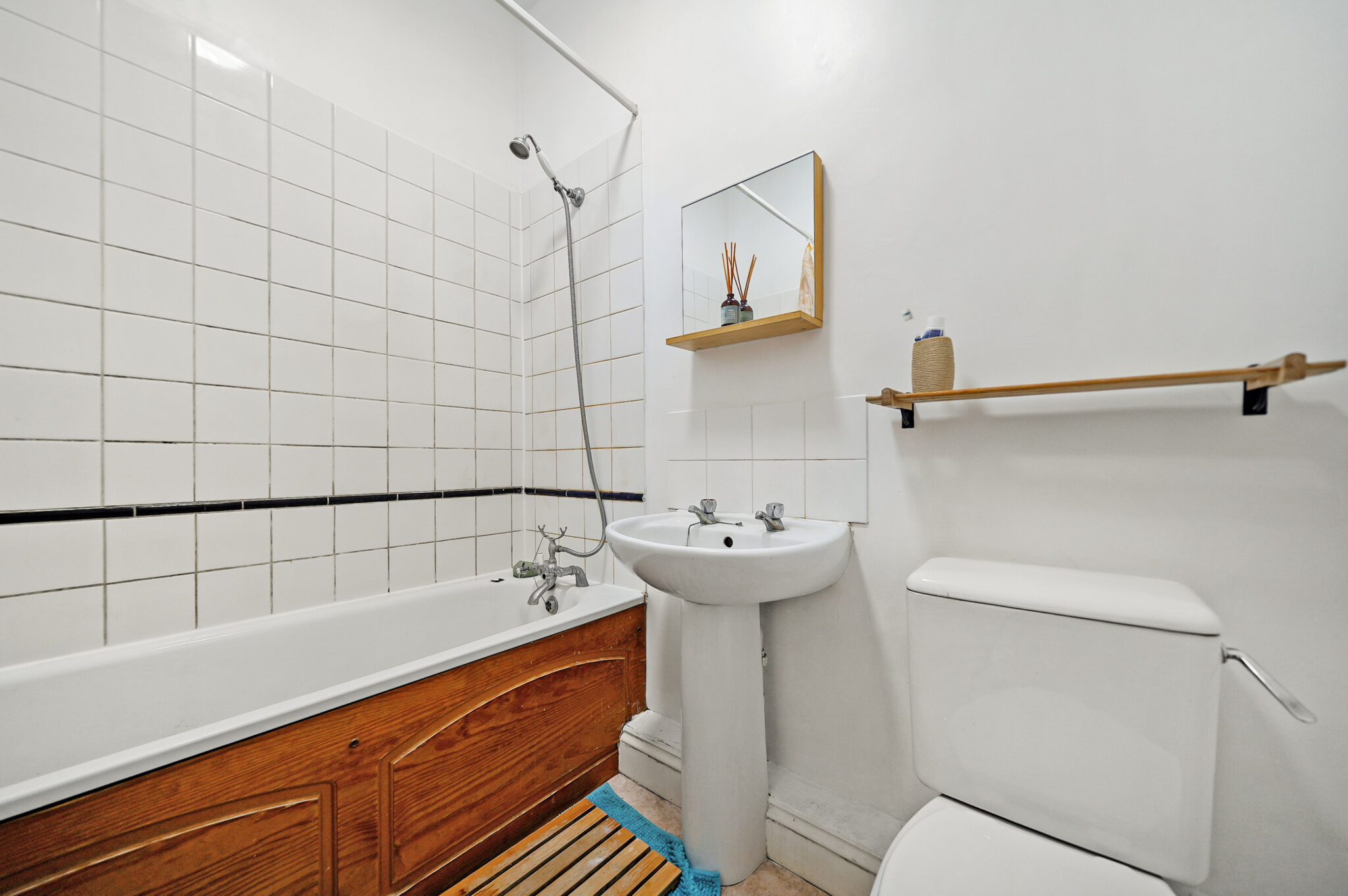 Compact bathroom with white tile walls, a wooden-front bathtub, pedestal sink, and a toilet. A wall mirror shelf and towel bar are also visible.