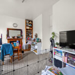 Messy living room with a pet/child gate in the foreground, a dining table cluttered with items and UK flags, and a large TV on the right. A tall shelf and wall clock are visible in the back.