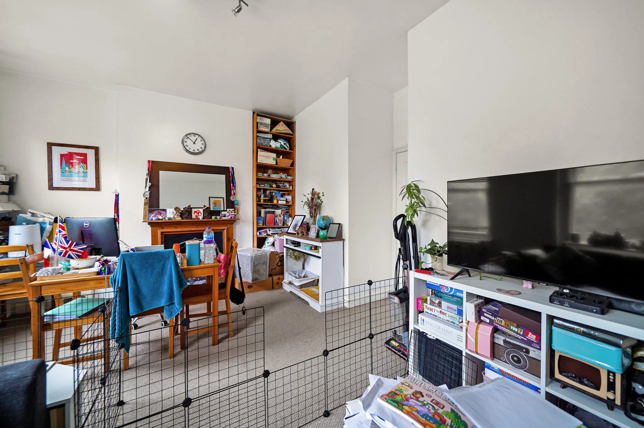 Messy living room with a pet/child gate in the foreground, a dining table cluttered with items and UK flags, and a large TV on the right. A tall shelf and wall clock are visible in the back.