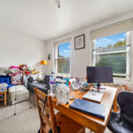 Messy living room and workspace with a wooden desk, laptop, water bottles, and clutter near a sofa by large windows.