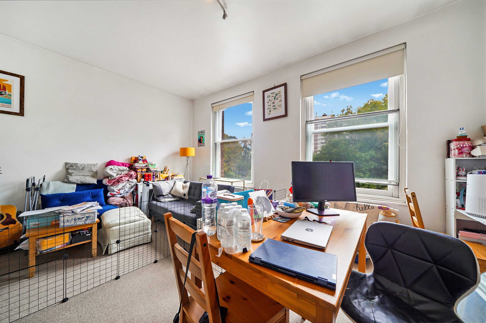 Messy living room and workspace with a wooden desk, laptop, water bottles, and clutter near a sofa by large windows.